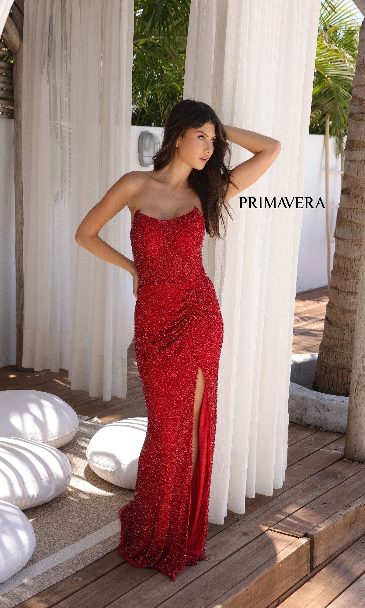 A woman wearing the Primavera 4364 long prom dress with a strapless neckline and thigh-high slit stands on a wooden deck beside white curtains and palm trees.